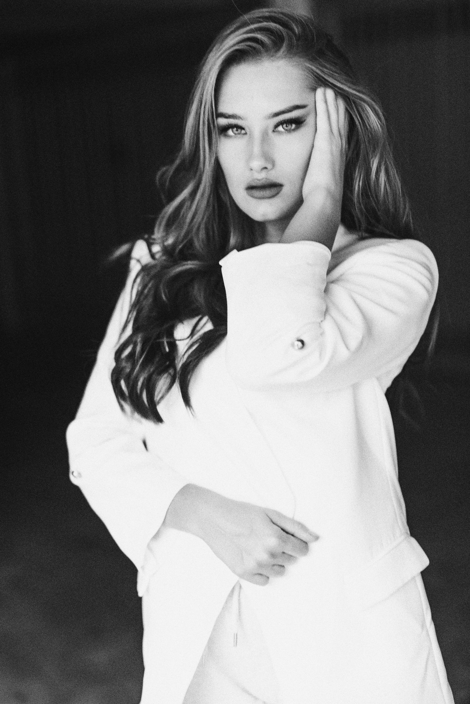 Posa a mezza figura in bianco e nero di una donna con giacca chiara, una mano che sfiora i capelli e lo sguardo fisso verso la fotocamera.