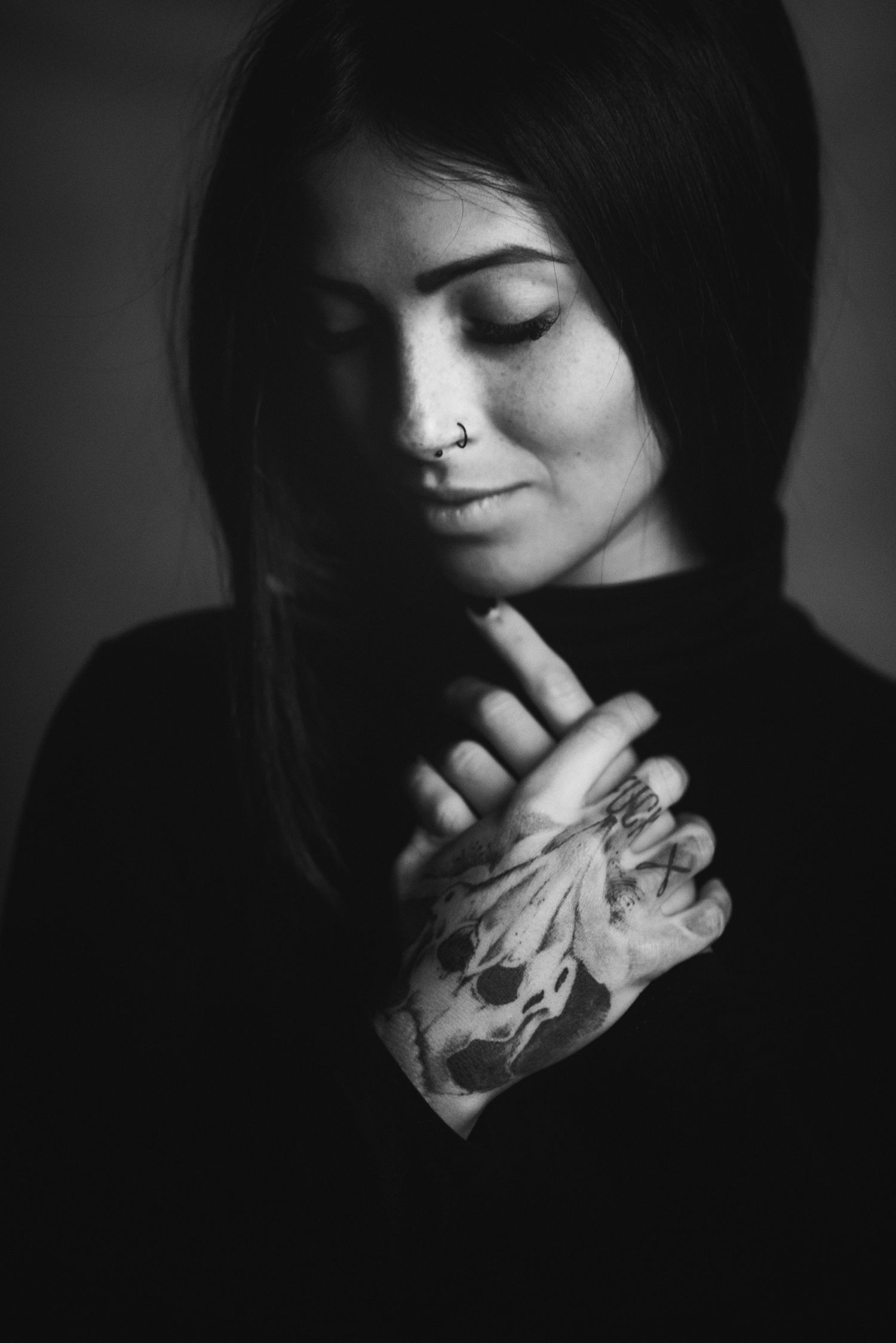 Primo piano intimo in bianco e nero di una donna con occhi socchiusi e piercing al naso, che tiene le mani tatuate con un teschio incrociate