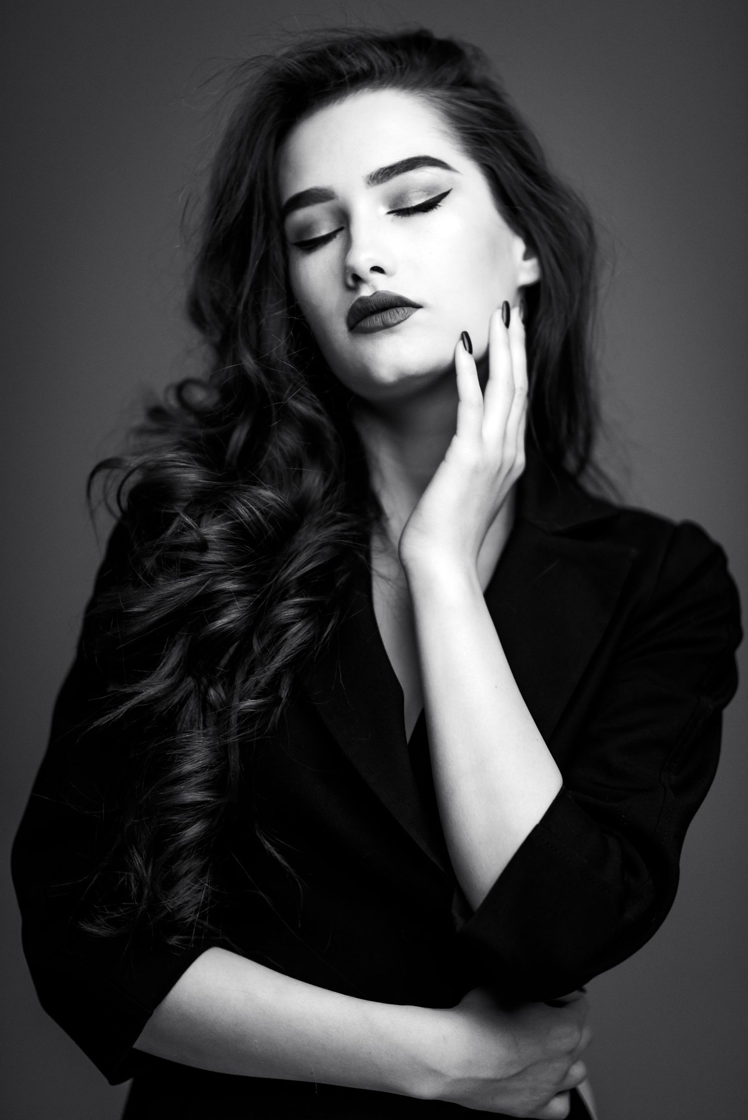 Ritratto glamour in bianco e nero di una donna con lunghi capelli mossi e occhi chiusi, con una mano appoggiata sulla guancia mentre indossa