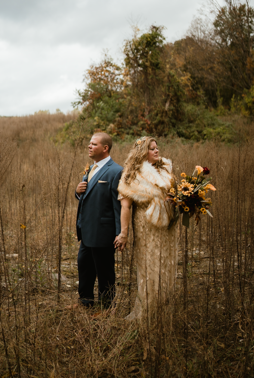 a married couple holding hands facing back to back looking out in the distance in a field during the fall