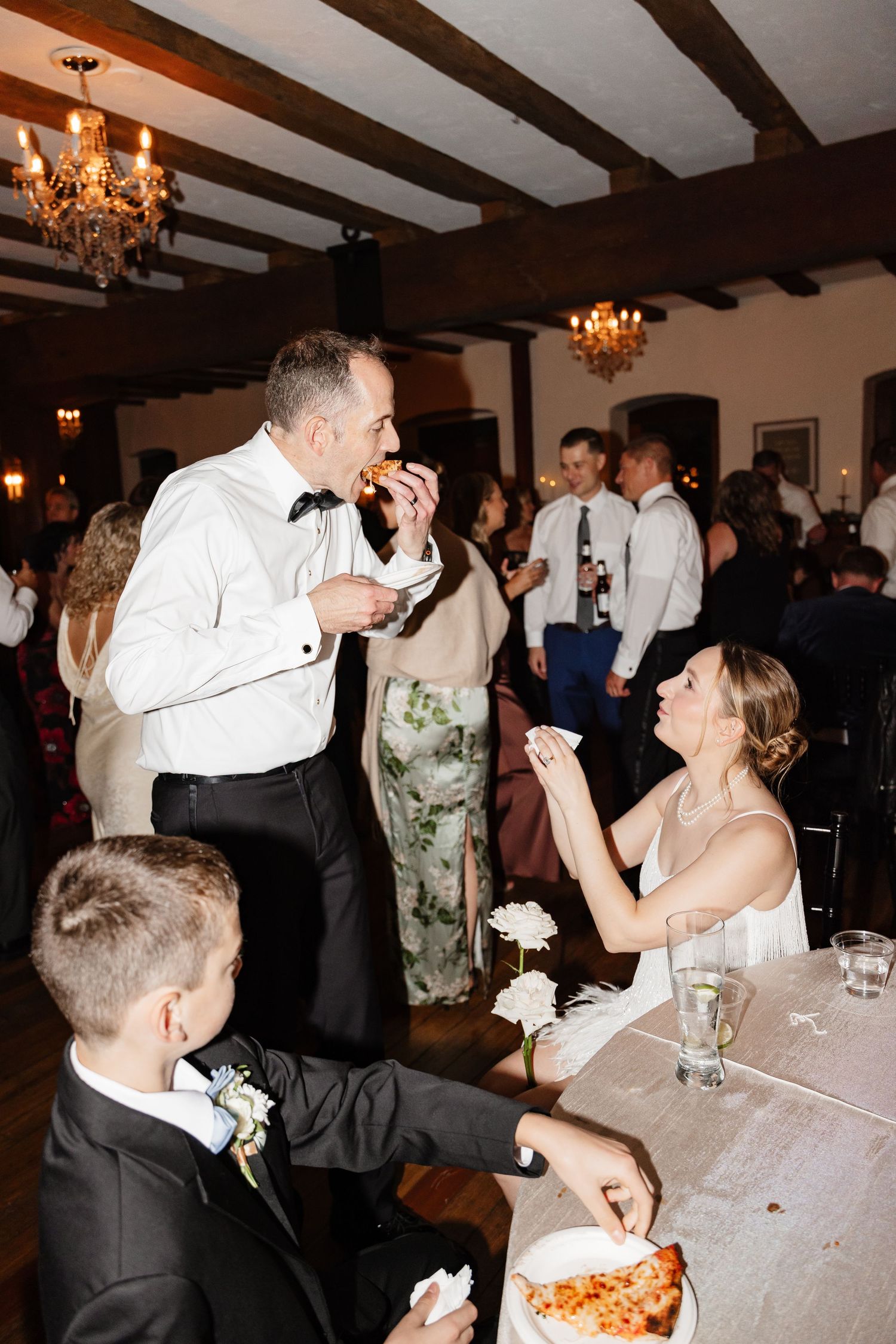 Groom in tuxedo eating pizza while bride laughs at wedding reception table in elegant ballroom.