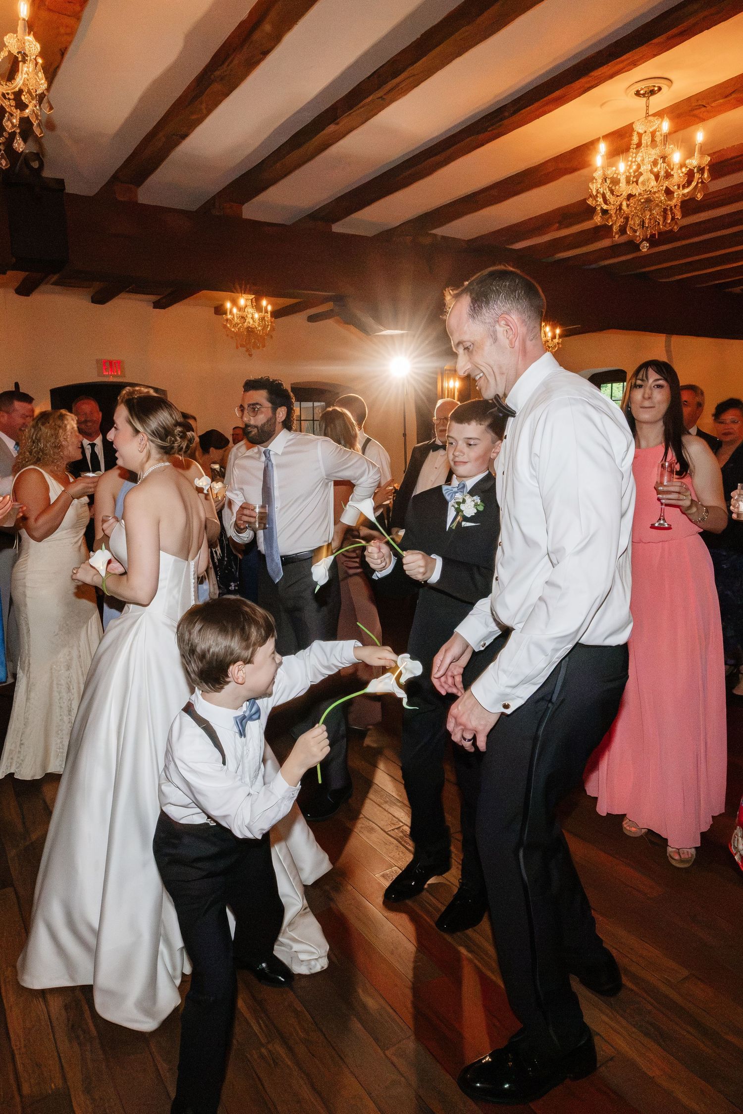 Wedding party dancing in rustic ballroom with chandeliers, children joining bride and groom on dance floor.