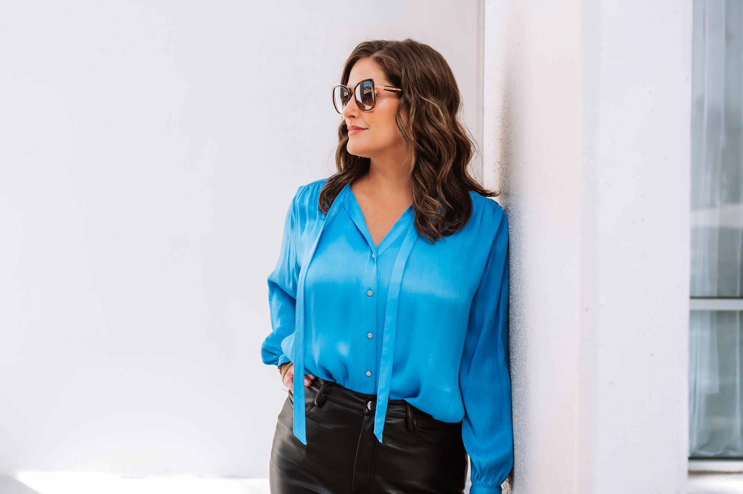 Stylish woman in blue satin blouse and sunglasses leaning against white wall outdoors.