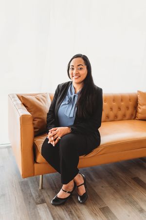 Professional Asian woman in black blazer sitting on tan leather sofa smiling.