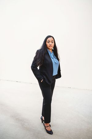 Confident woman in black business suit and blue blouse posing against white wall.