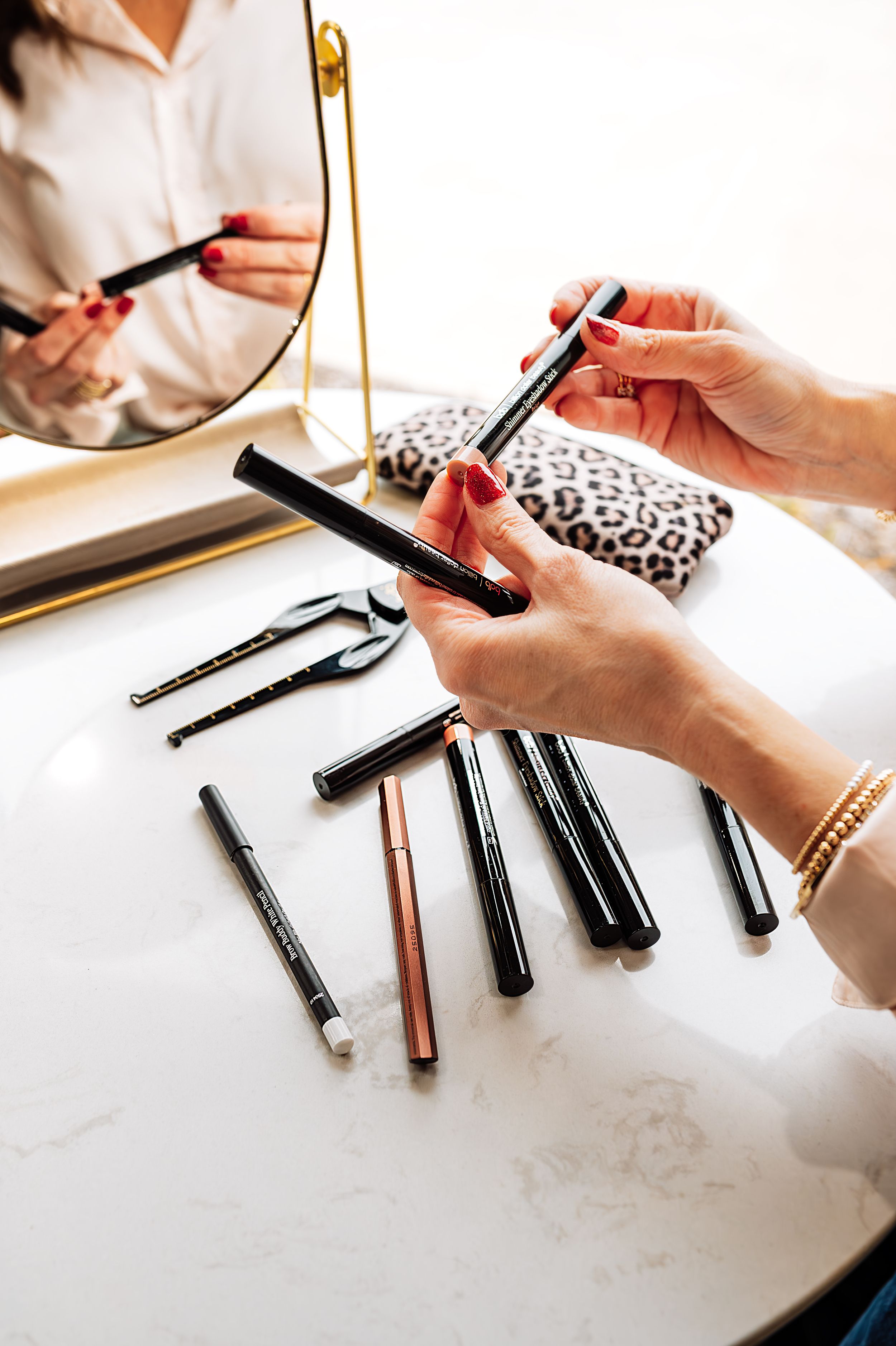Close-up of hands holding makeup pencils near a mirror with various cosmetics on a marble surface.