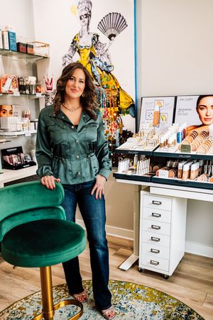 Woman in green jacket stands beside velvet stool in beauty boutique with makeup displays.
