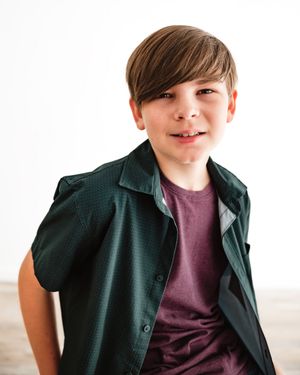 Young boy smiling in dark green shirt over maroon tee on white background.