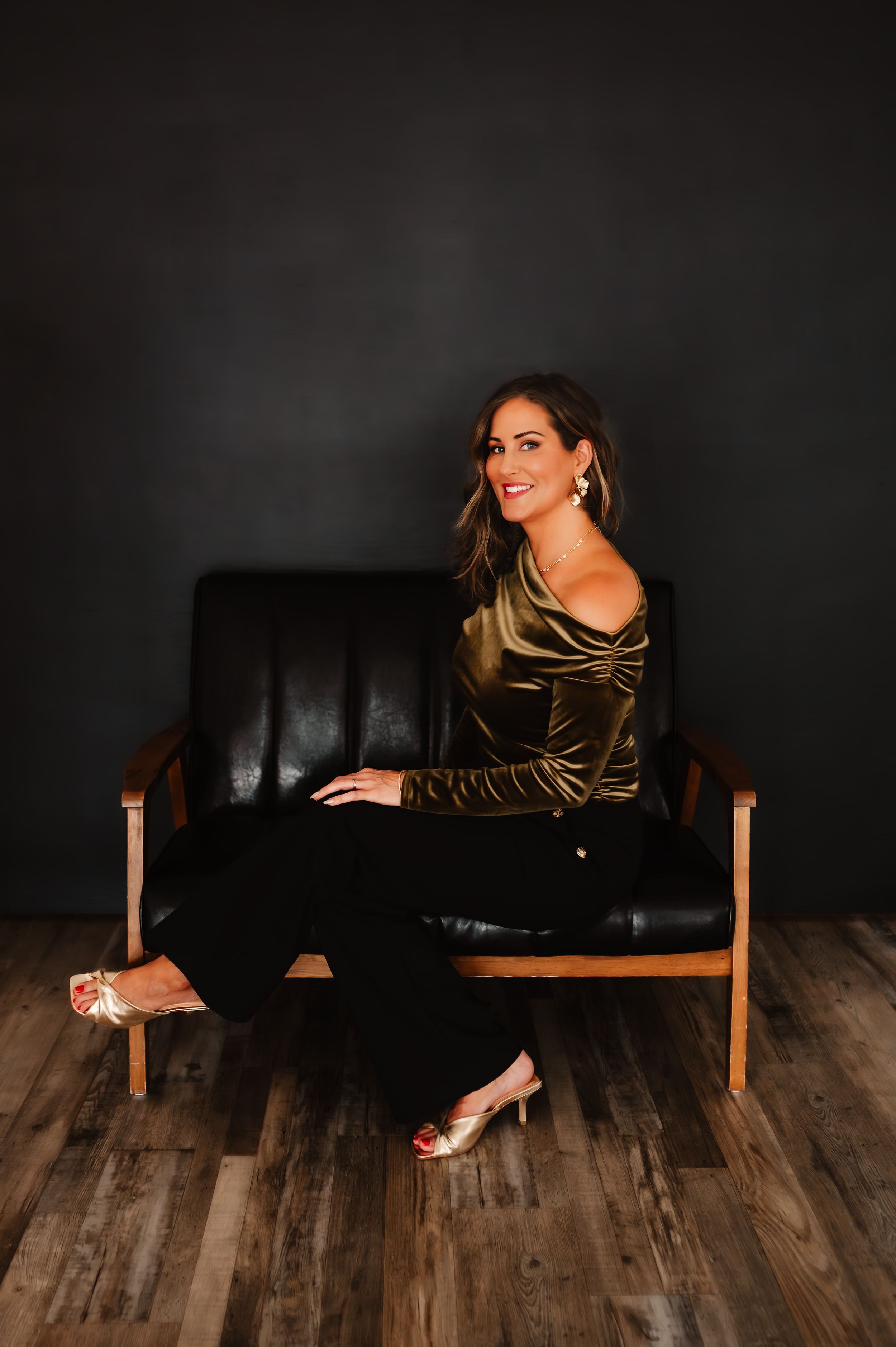 Woman in gold velvet top sits relaxed on black leather chair against dark wall.