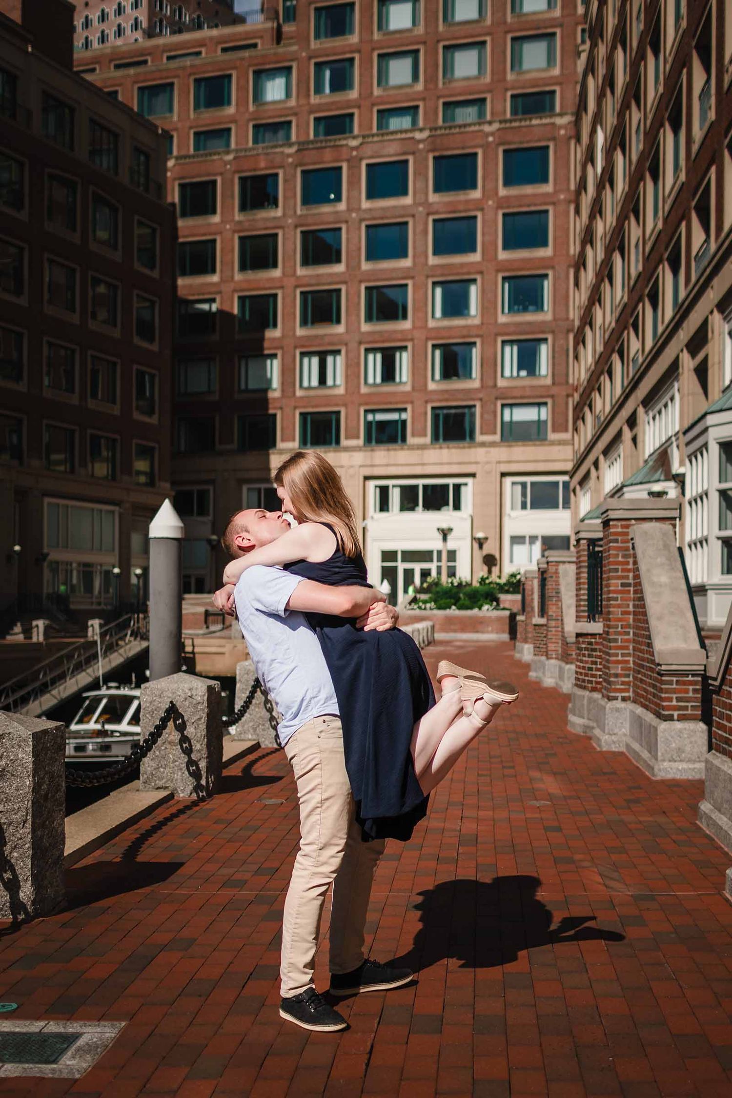 Groom lifting his fiancée for a kiss during their engagement session near Rowes Wharf in Boston