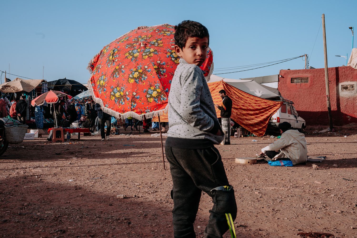 Young boy stands in a dusty refugee camp holding a colorful red floral umbrella amid tents and people.