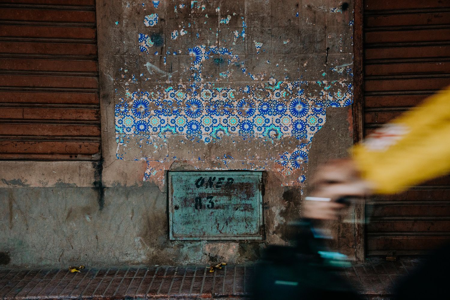 Weathered wall with blue floral tile mosaic, rusty shutters, and a blurred cyclist passing by on a street.