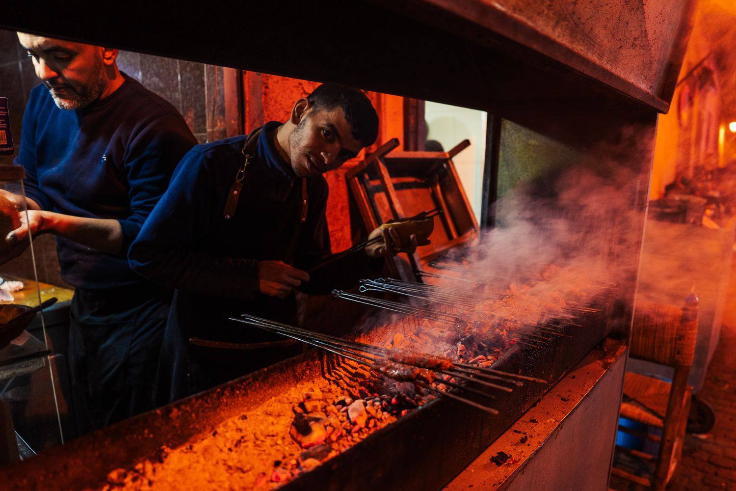 Two chefs grilling skewers over glowing charcoal embers at a smoky outdoor BBQ grill at night.