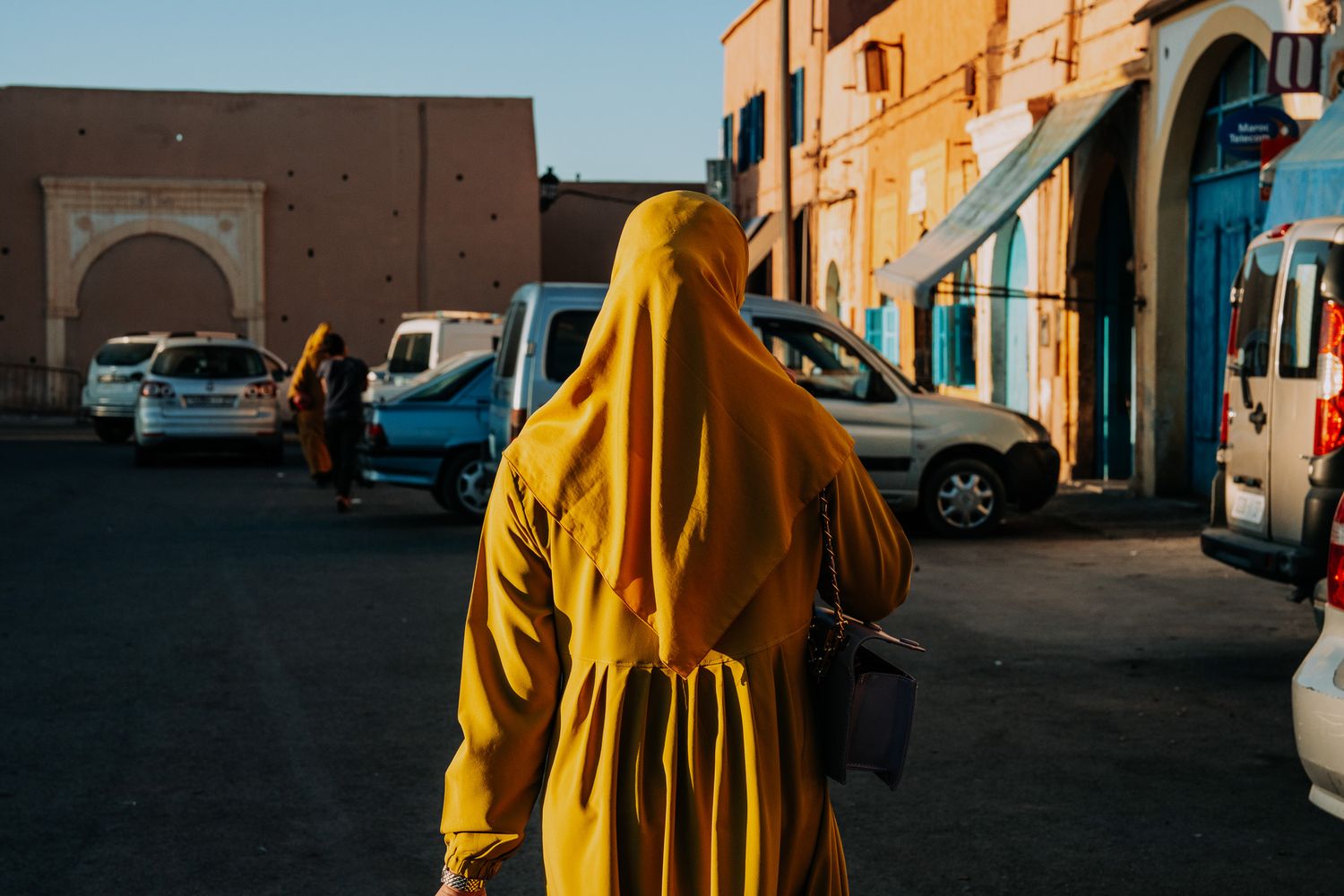 A woman in a vibrant yellow hijab walks through a sunlit Moroccan street with cars and traditional buildings.