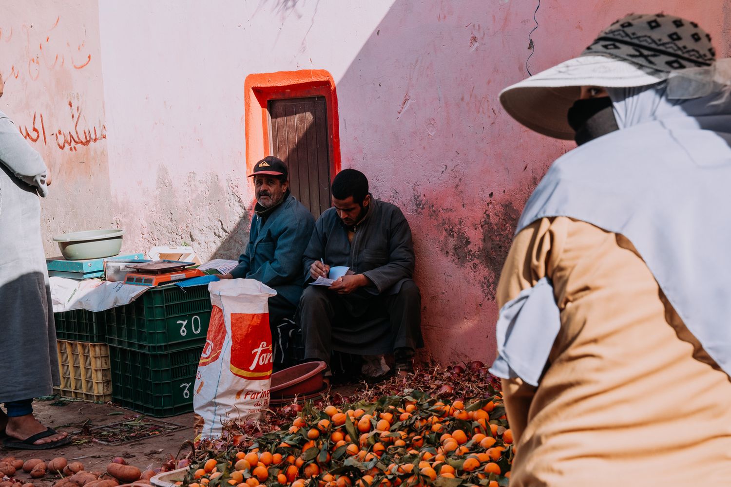 Moroccan street vendors selling fresh oranges at a colorful market stall against a red and pink wall.