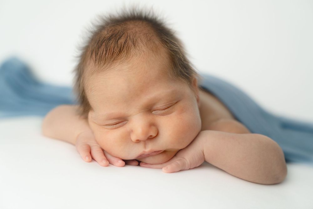 Peaceful newborn baby sleeping with hands under chin, wrapped in a soft blue blanket on a white background.
