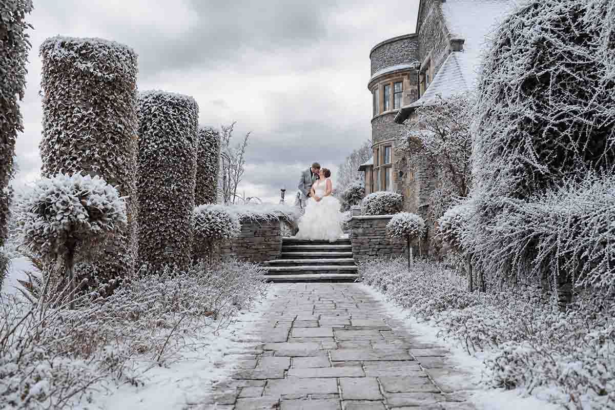 Bride and groom kiss on snowy castle steps surrounded by frost-covered hedges and trees in a winter wonderland wedding.