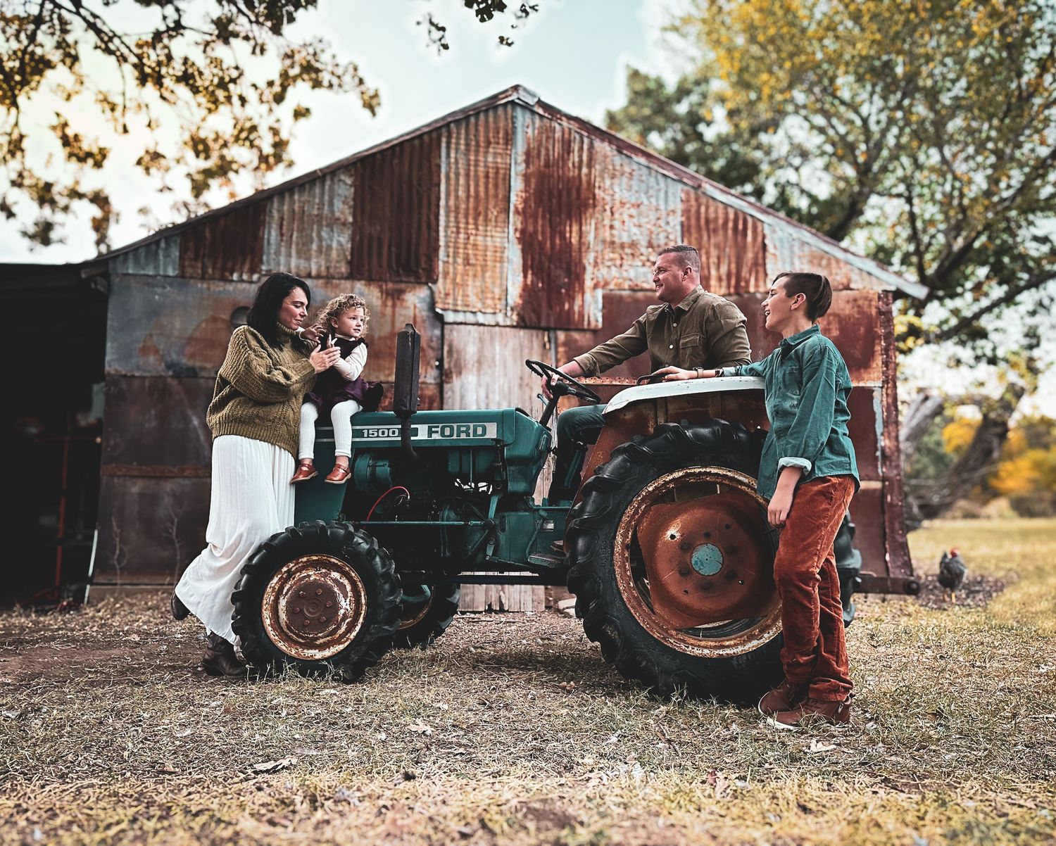 Family of four laughing together around a vintage green tractor near a rustic barn in autumn.