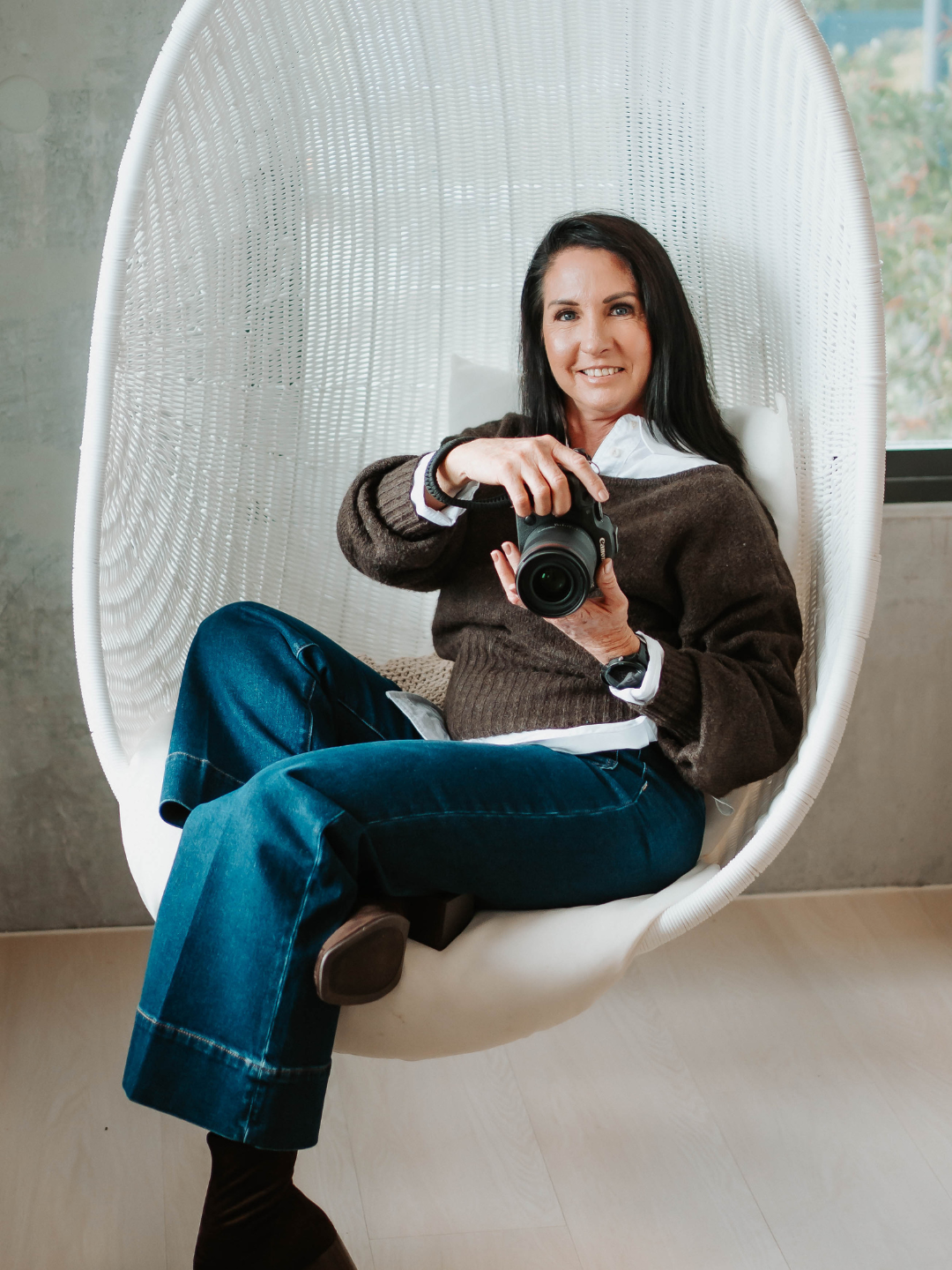 Woman sitting in white egg chair holding a DSLR camera, wearing brown sweater and blue jeans, smiling at camera.