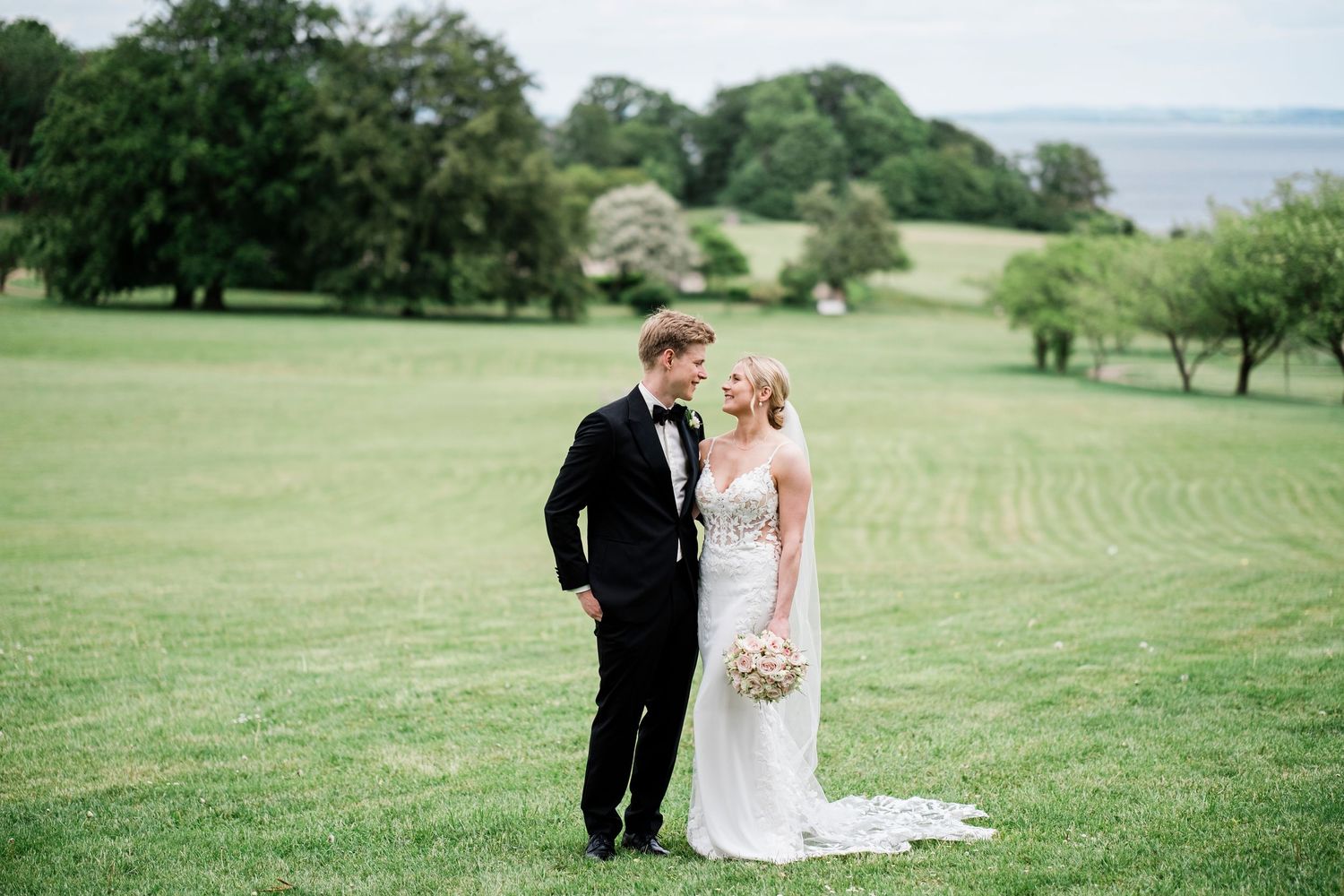 Bride and groom standing together on lush green lawn with scenic water view on their wedding day.