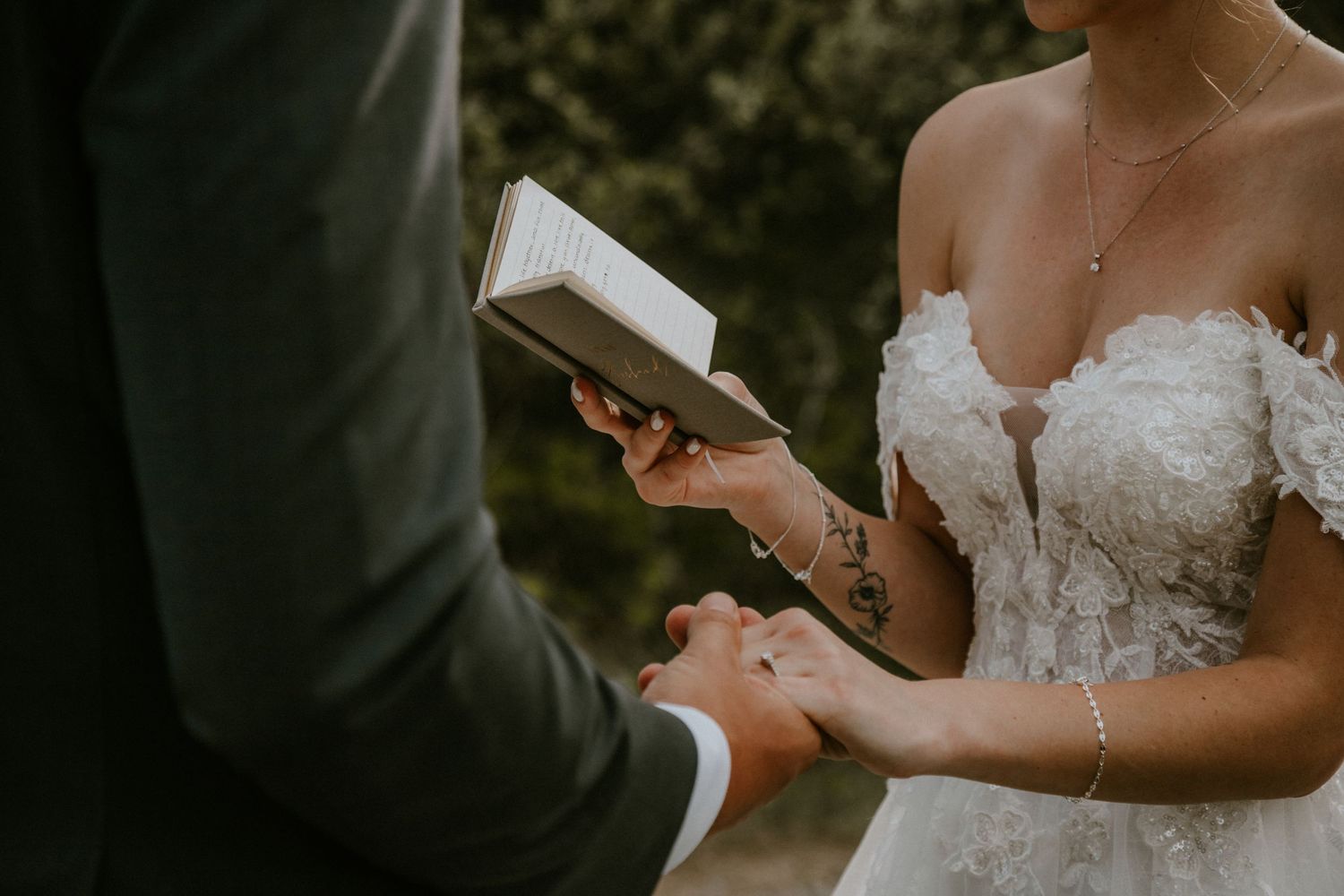 Bride in floral off-shoulder gown holds hands with groom during outdoor wedding ceremony vow exchange.