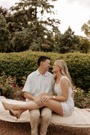 Couple sitting together on a stone bench in a lush garden, smiling at each other during an engagement photo session.