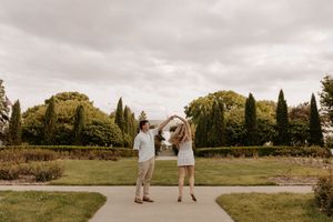 Couple dancing on garden path with cypress trees and cloudy sky backdrop during outdoor engagement session.