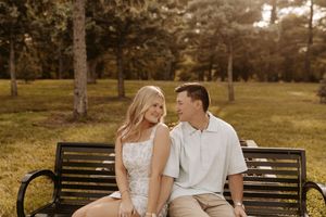 Couple sitting on a park bench smiling at each other, surrounded by lush green trees in warm golden light.
