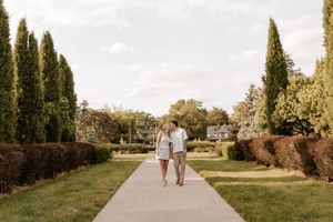 Couple walking hand in hand along a garden pathway lined with tall cypress trees and manicured hedges.