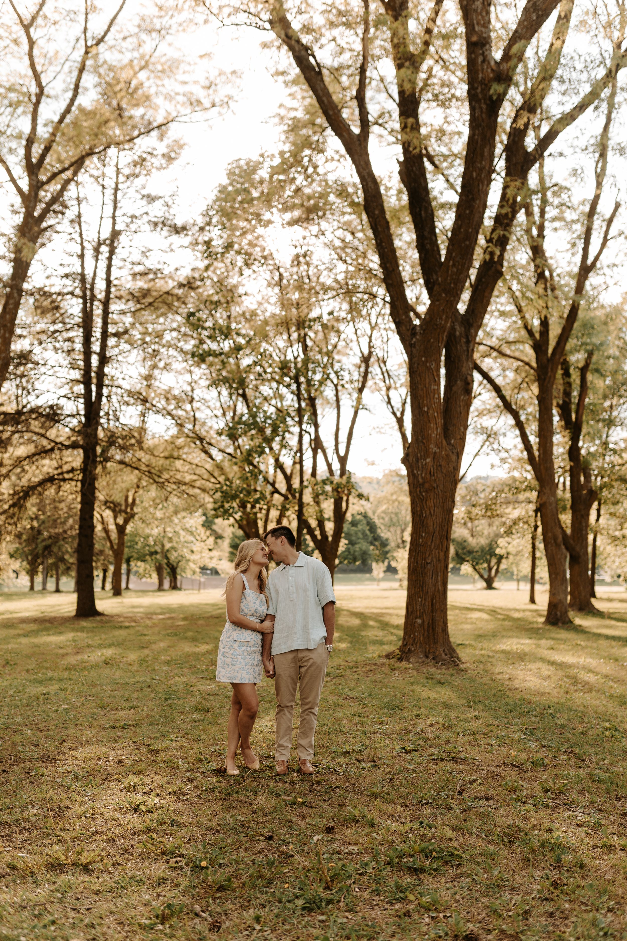 A couple shares a romantic moment standing together in a sunlit park surrounded by tall trees and lush green grass.