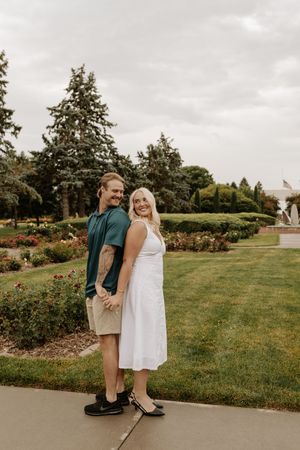 Couple holding hands in a lush park, woman in white dress and man in green shirt, smiling outdoors.