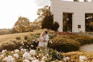 Couple embracing among white flowers and lush gardens near a white building at golden hour.