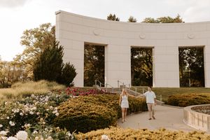 Couple walking hand in hand along a garden path near an elegant curved white building during an outdoor engagement shoot.