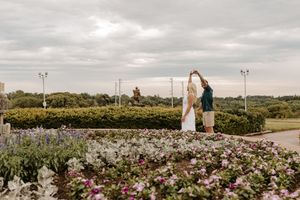 Couple dancing in a flower garden with lush greenery and a statue in the background under a cloudy sky.