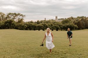 A bride in a white dress and groom in green walk across a lush green field with trees and a stone tower backdrop.