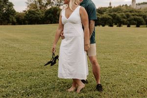 Pregnant woman in white dress holding heels walks barefoot on green grass with partner during outdoor maternity photoshoot.