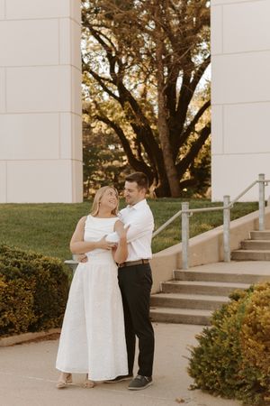 Couple in white dress and white shirt embracing and laughing outdoors near stone building steps and autumn trees.