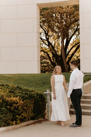 Couple holding hands in front of a stone building archway with golden trees, woman in white midi dress and man in white shirt.