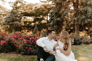 A couple in wedding attire sits together on grass, gazing lovingly at each other with blooming flowers and trees behind.