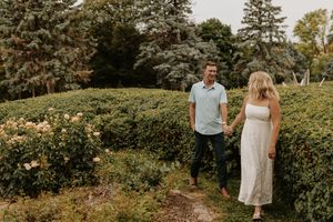 Couple holds hands walking through lush garden hedges with blooming flowers and tall trees in background.