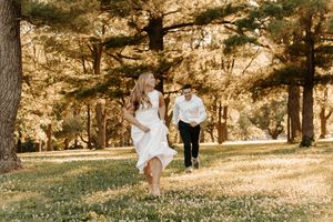 Couple in white outfits running through a sunlit park with tall trees and green grass during an outdoor engagement session.