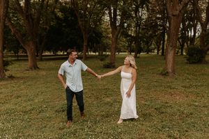Couple holding hands in a lush green park, man in blue shirt and woman in white dress, surrounded by trees.