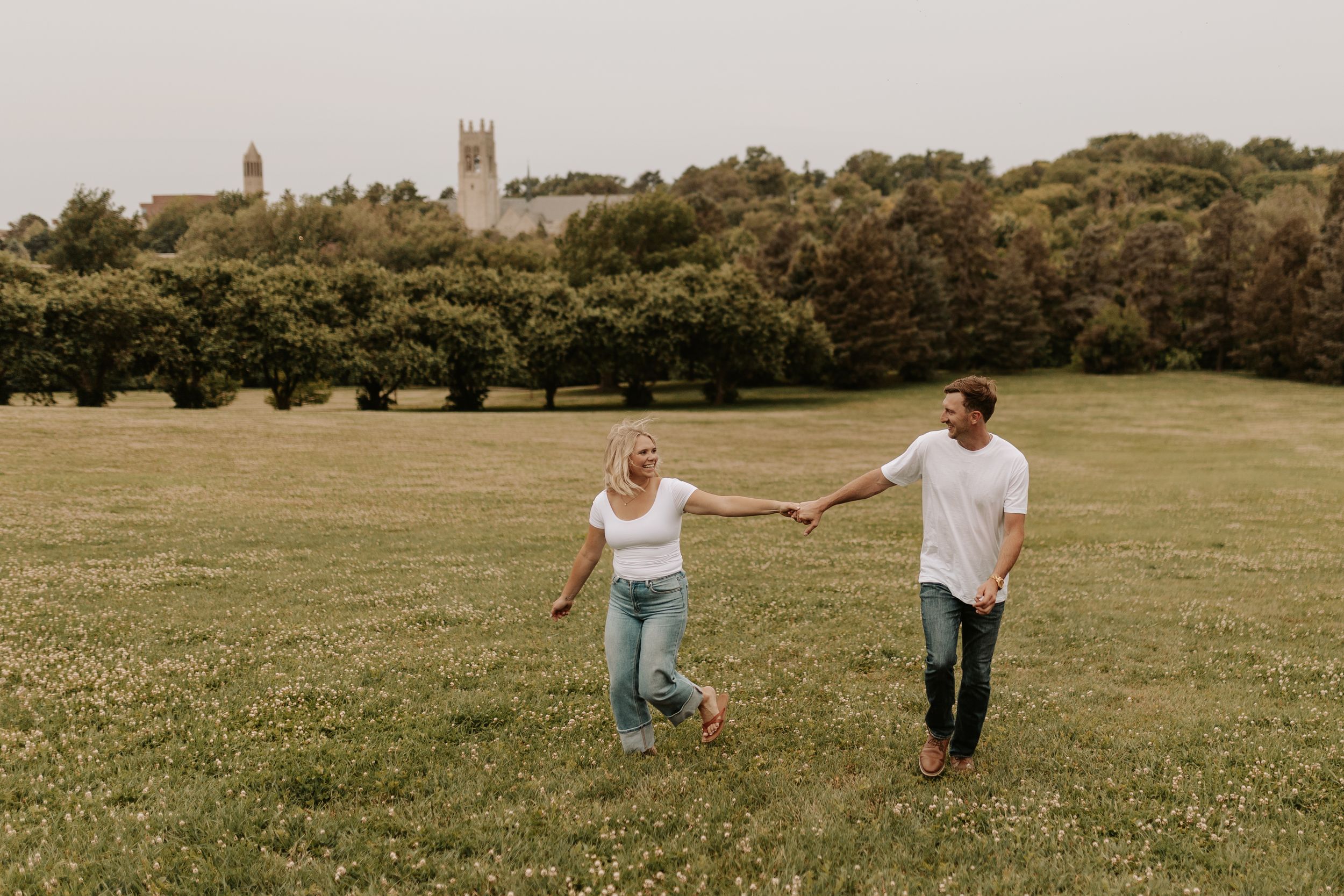 A couple holds hands and walks through a green field with trees and a historic stone building in the background.