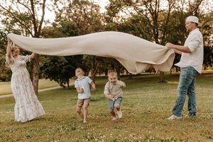 Family of four playing with a large blanket outdoors in a lush green park at golden hour.