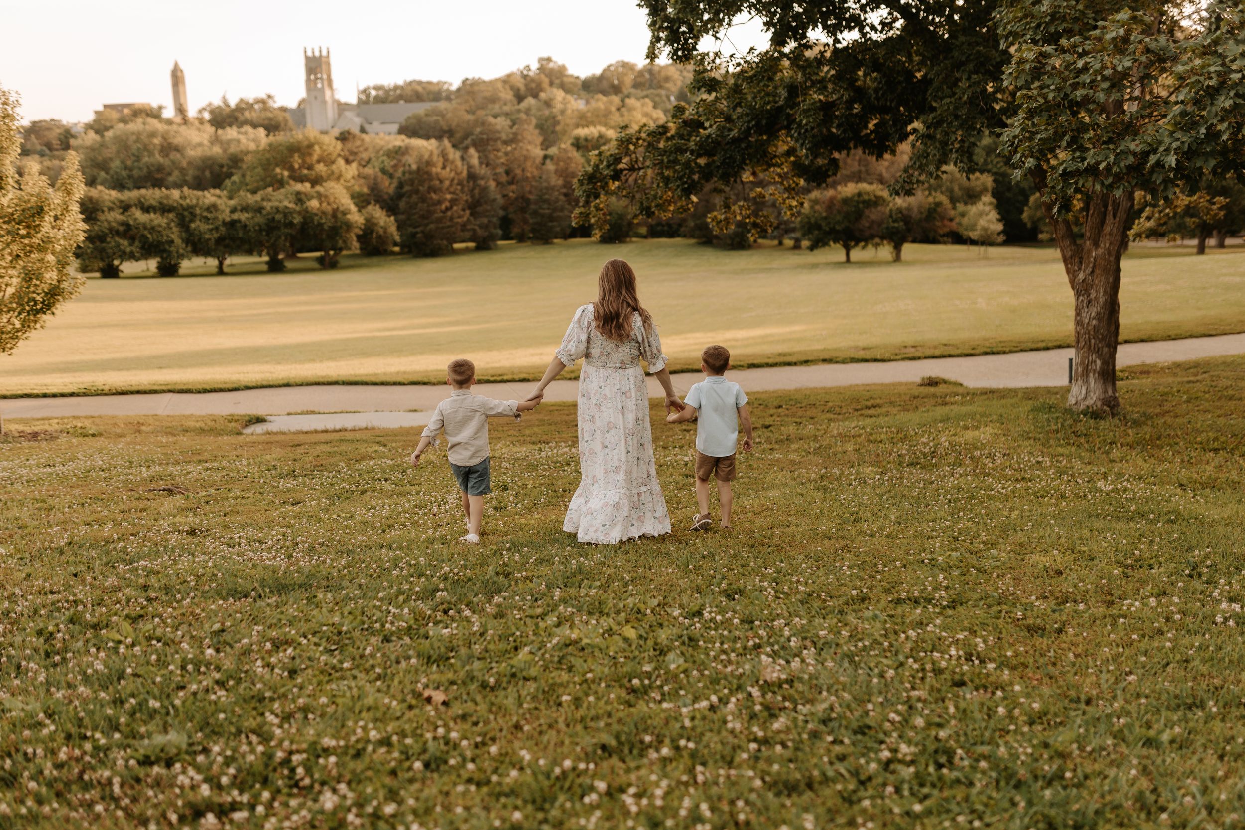A bride in a white gown walks hand-in-hand with two young boys across a lush green park landscape.