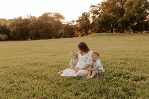 A pregnant mother sits on lush green grass at golden hour with two young children leaning against her.