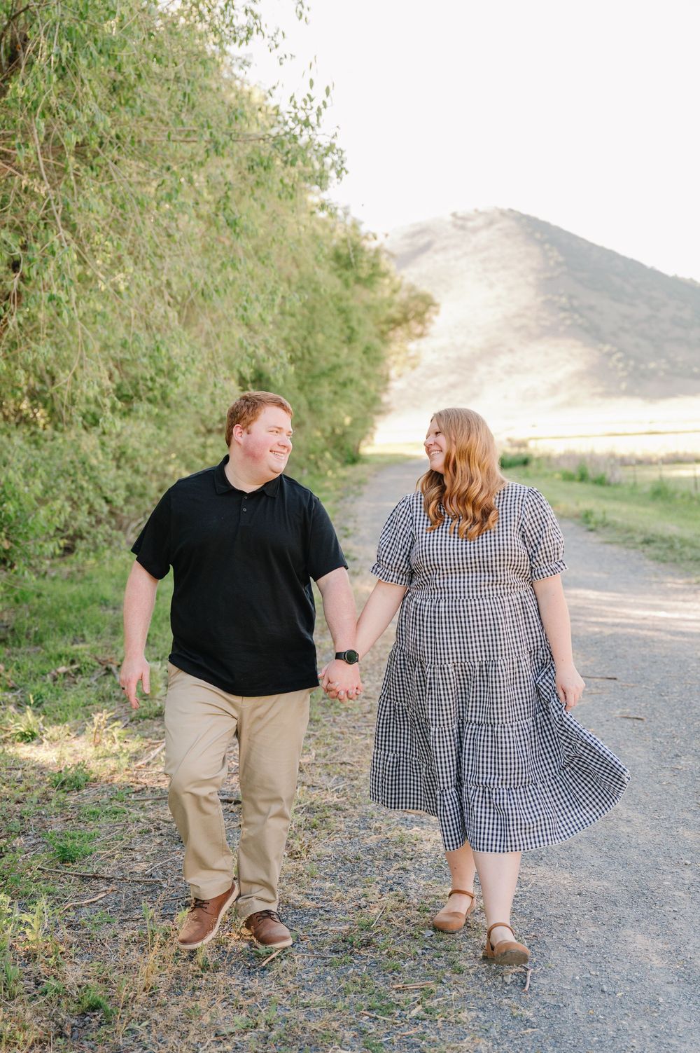 A couple with red hair holds hands while walking on a dirt path, surrounded by greenery and a hill backdrop.