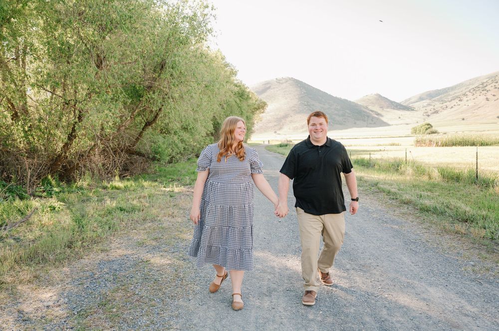 Couple holding hands walking on a gravel path with green trees and rolling hills in the background.