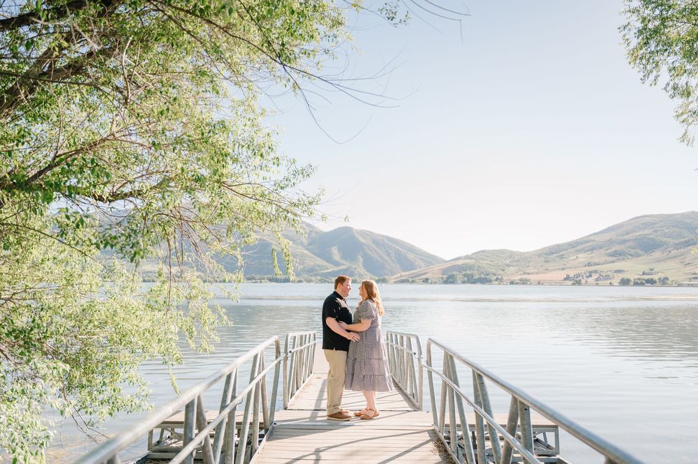 Couple embracing on a wooden dock overlooking a serene mountain lake surrounded by lush green trees on a sunny day.