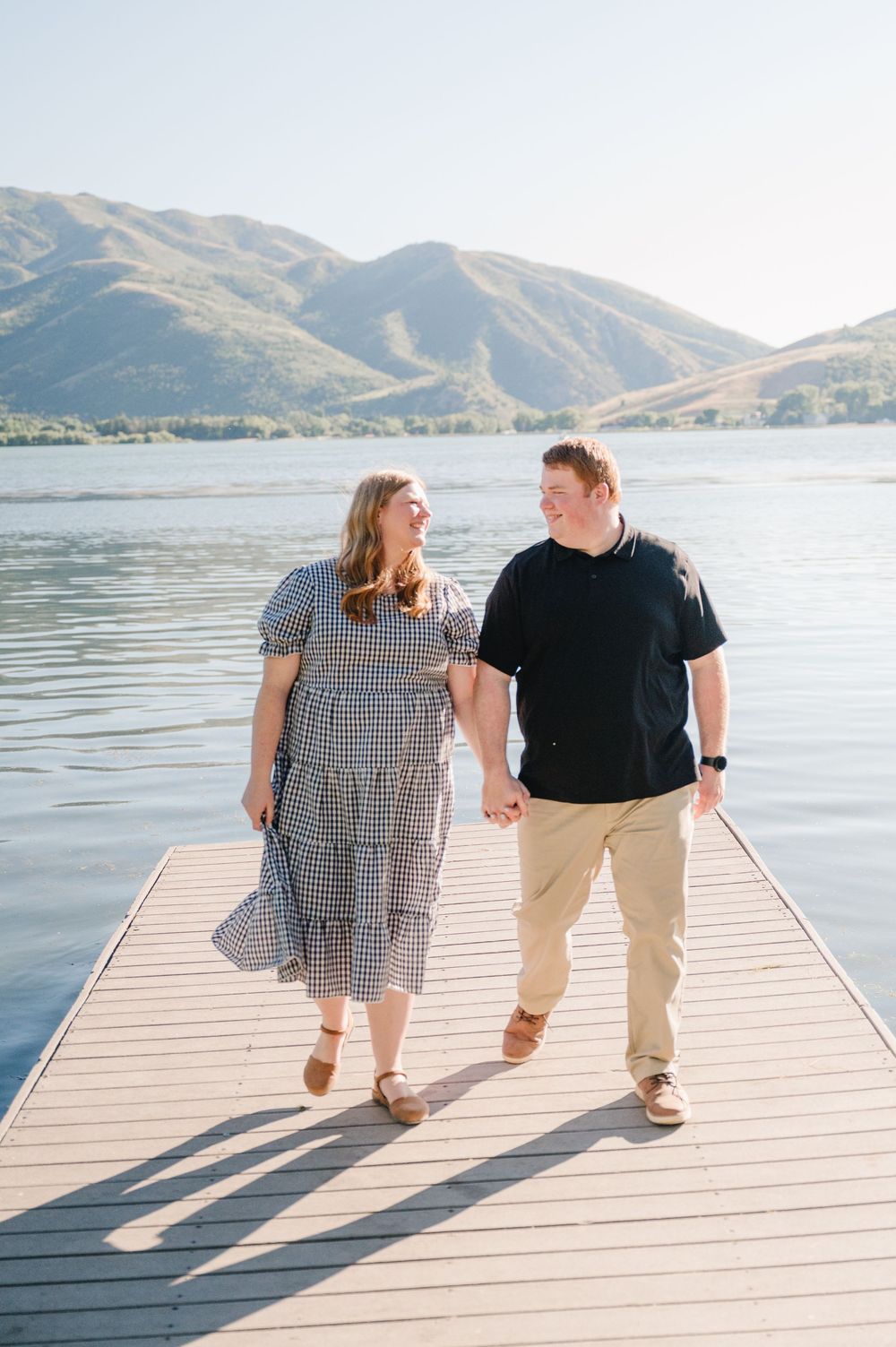 Couple holding hands and walking on a dock with a lake and mountains in the background on a sunny day.